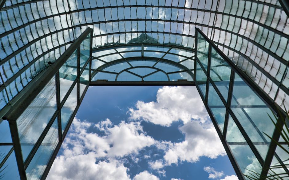 Glass arched roof of a greenhouse against a blue sky with clouds, showcasing modern architecture.