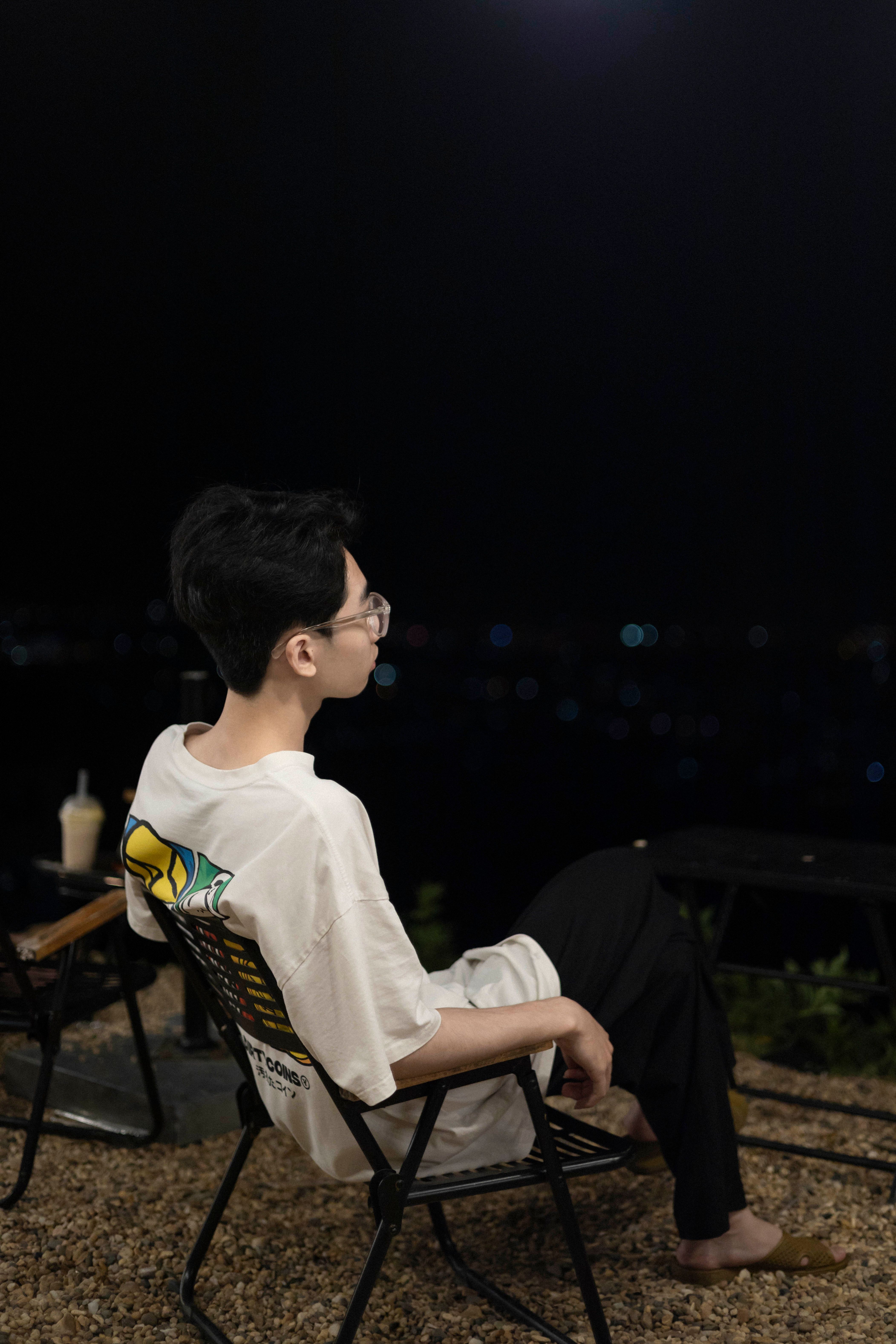 A young man sits on a chair outdoors at night, enjoying a peaceful moment.