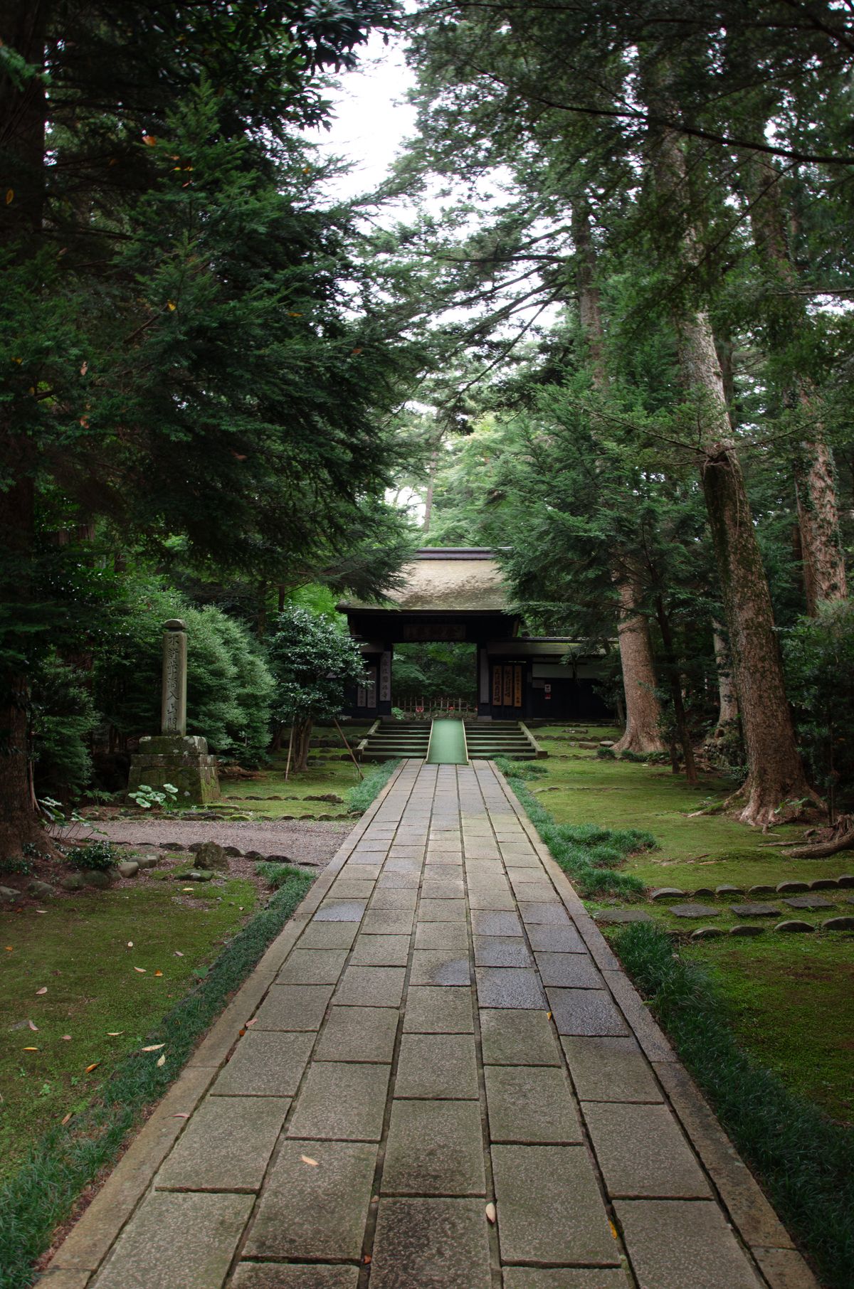 A tranquil stone walkway leading through lush forest toward a traditional wooden building in Japan