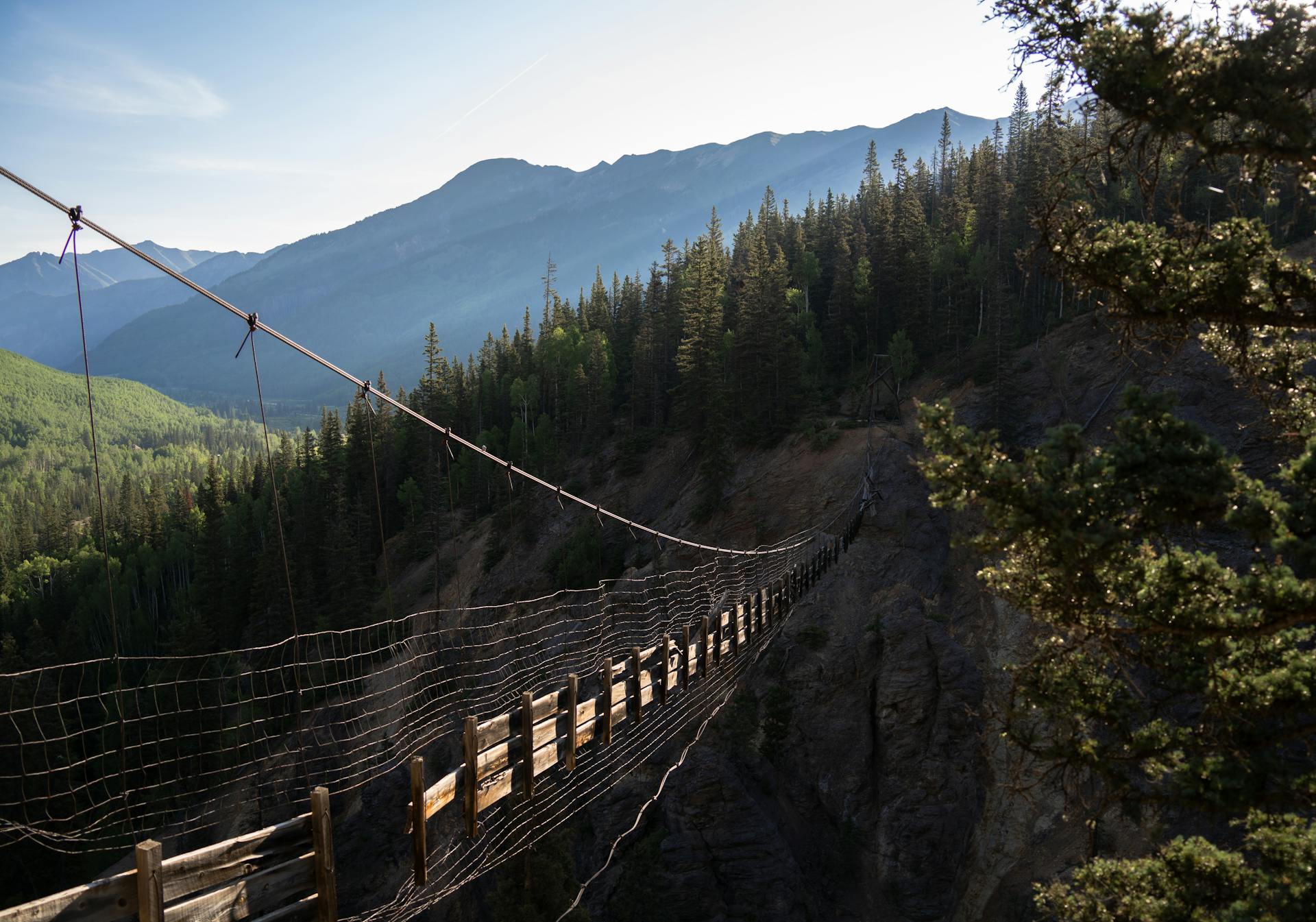 A wooden suspension bridge spans a lush forested valley with towering mountains in the background.