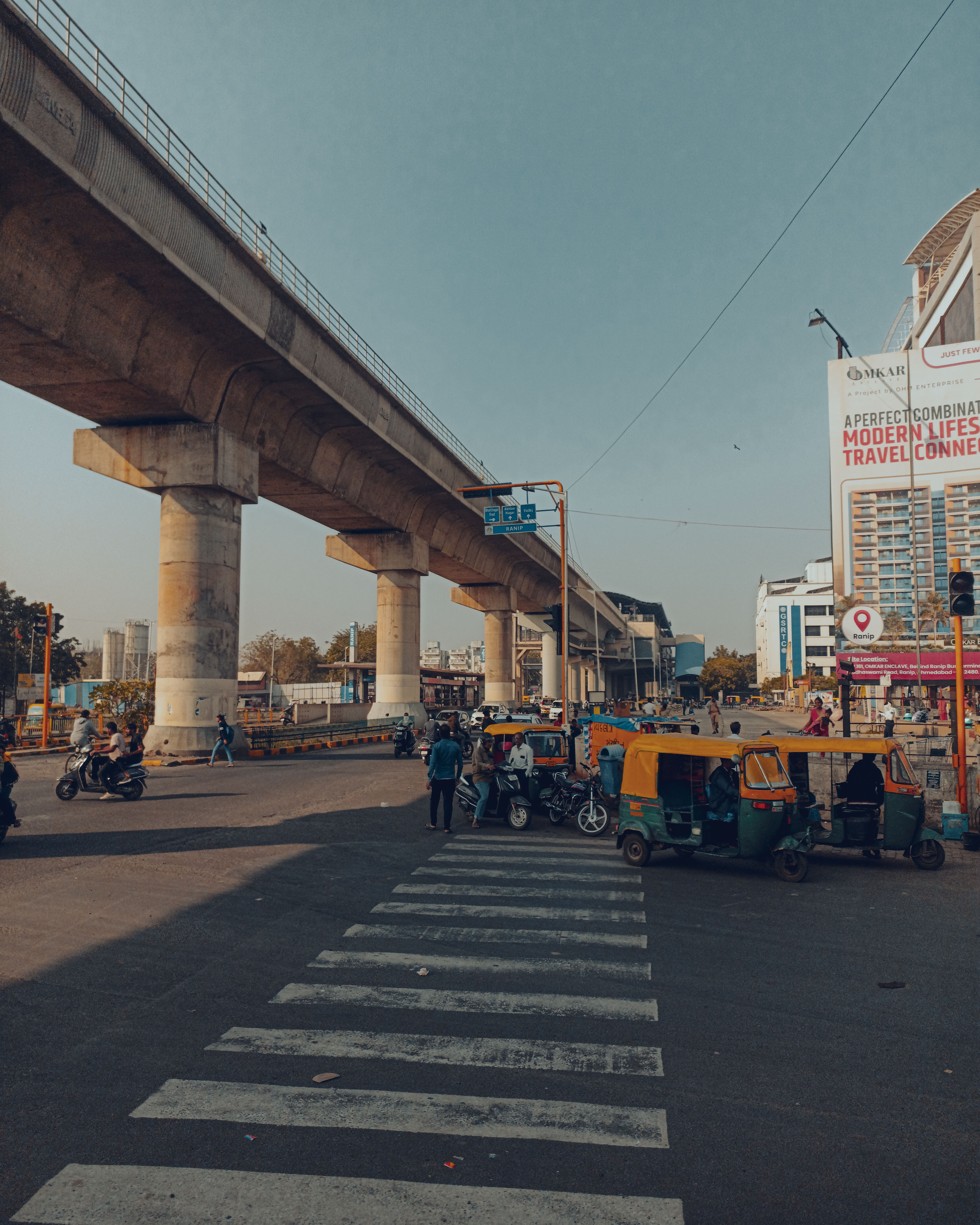 A busy urban intersection in India featuring an elevated metro line with rickshaws and pedestrians.