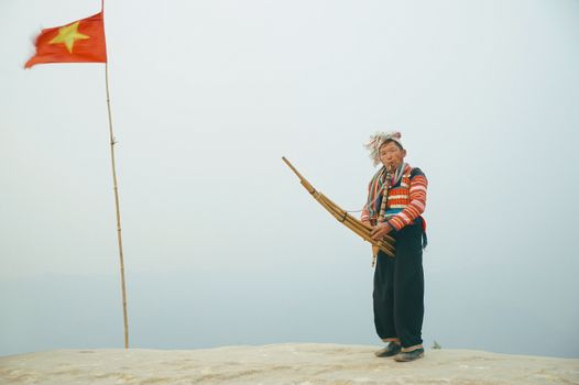 A traditional Vietnamese musician playing a bamboo instrument under the national flag. Captured in Sơn La, Việt Nam.