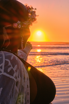 A woman wearing a floral crown gazes at the sunset over the ocean, capturing a serene beach moment.