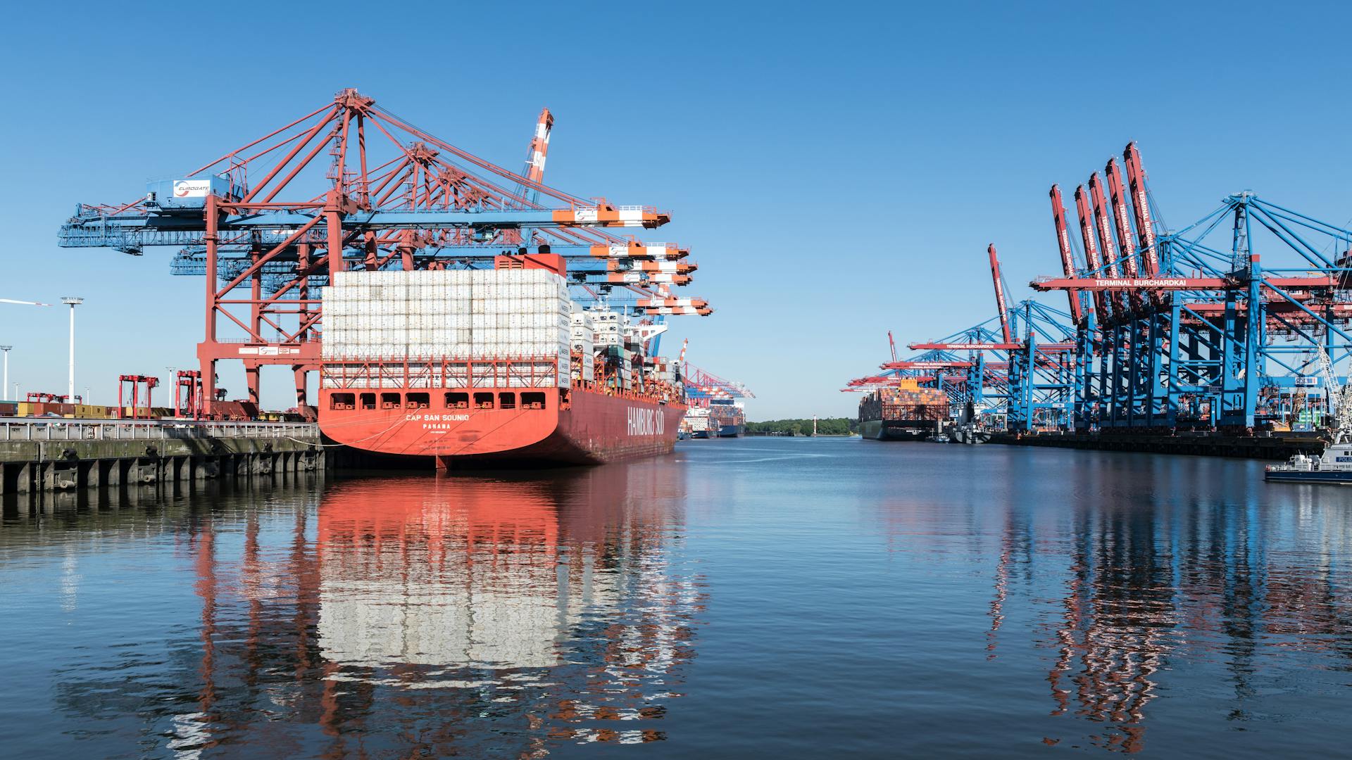 Vibrant container ship docked at Hamburg harbor, reflecting on calm water under a clear blue sky.