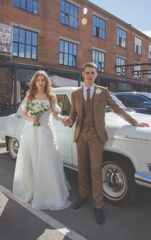 Bride and groom posing by a vintage car on a sunny day in the city.