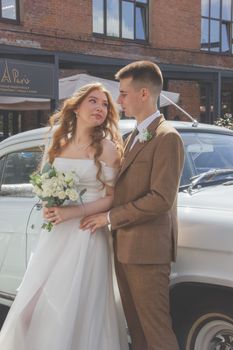 Beautiful bride and groom posing by a classic car outdoors on their wedding day.