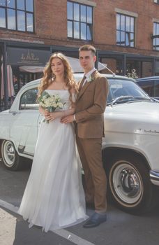 Bride and groom pose beside a classic car on their wedding day, capturing a timeless moment.