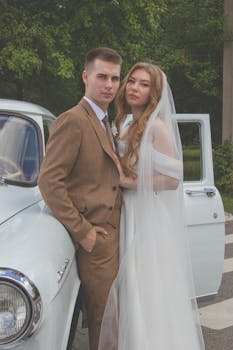 A stylish couple in wedding attire leaning on a classic vintage car outdoors.