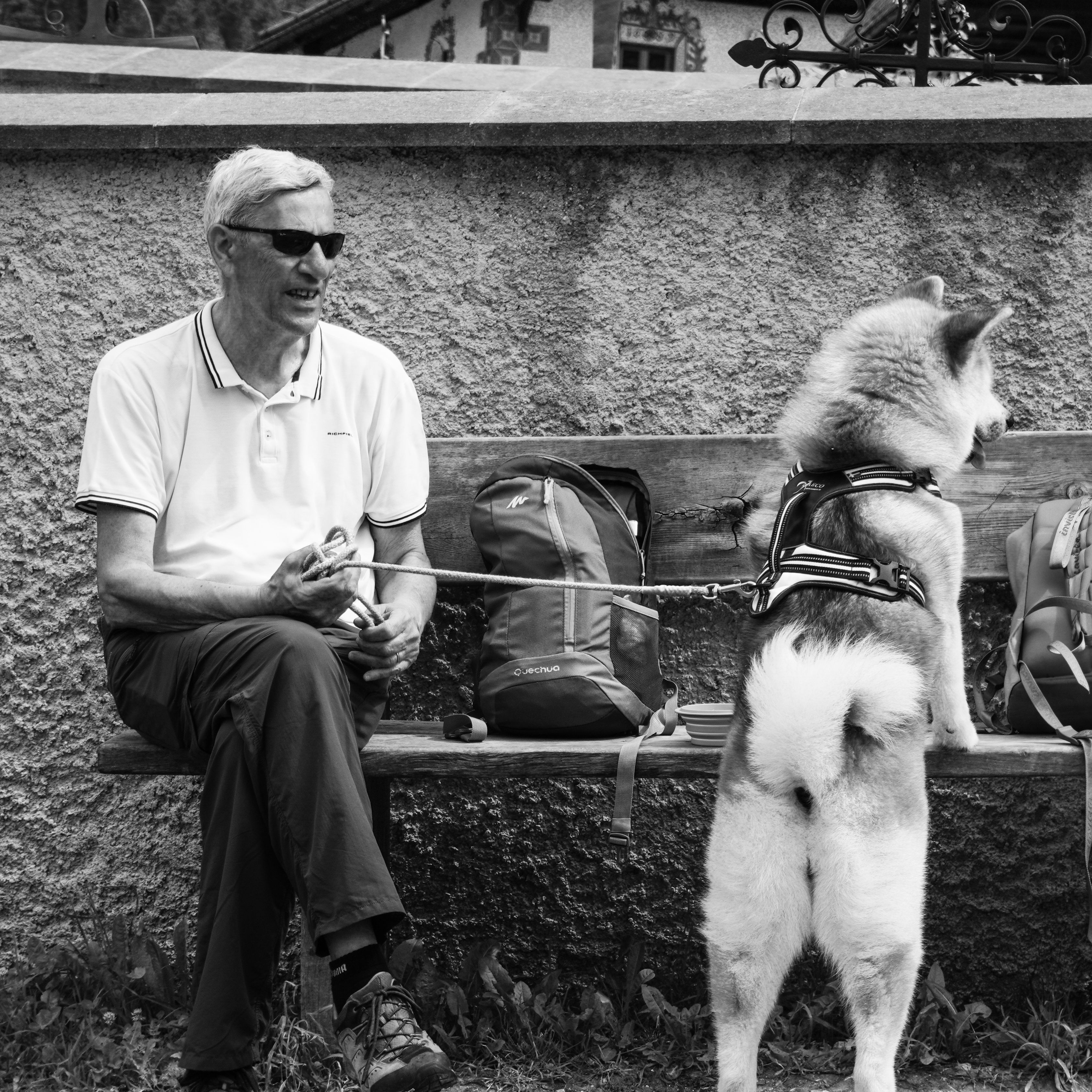 Black and white image of a senior man sitting with a dog beside him on an outdoor bench.