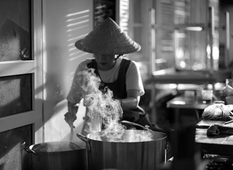 A street food vendor wearing a traditional hat cooking on a stove indoors, captured in moody monochrome.