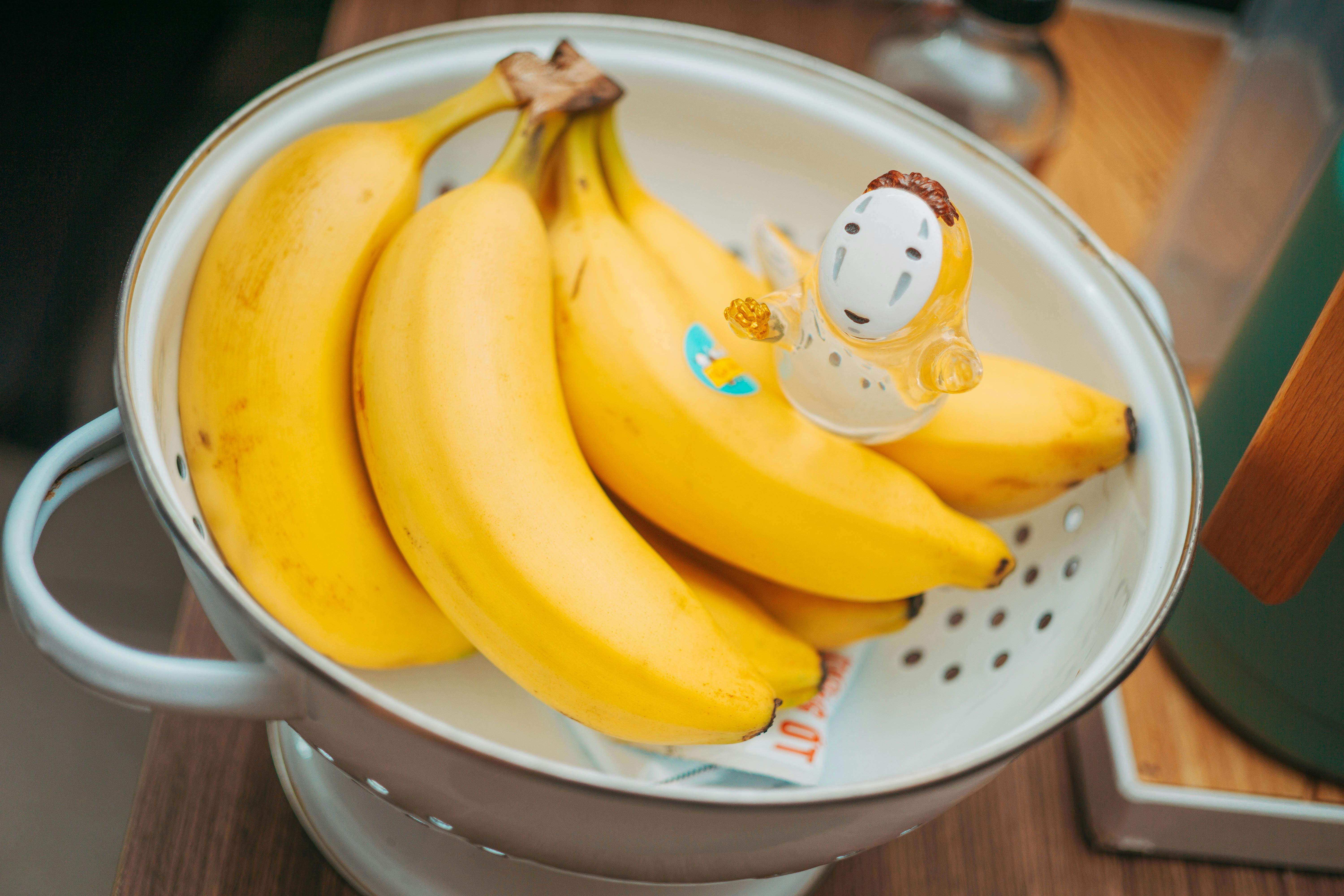 Three ripe bananas in a metal colander with a small toy figure, on a wooden table.