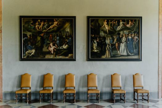 A beautifully designed interior with ornate paintings and classic wooden chairs lined up against the wall.
