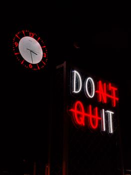 Neon sign reading 'Don't Quit' beside a wall clock at midnight. Encouragement theme in urban setting.