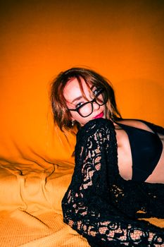 A stylish woman poses in black lingerie, wearing glasses and a lace robe against a vibrant background.