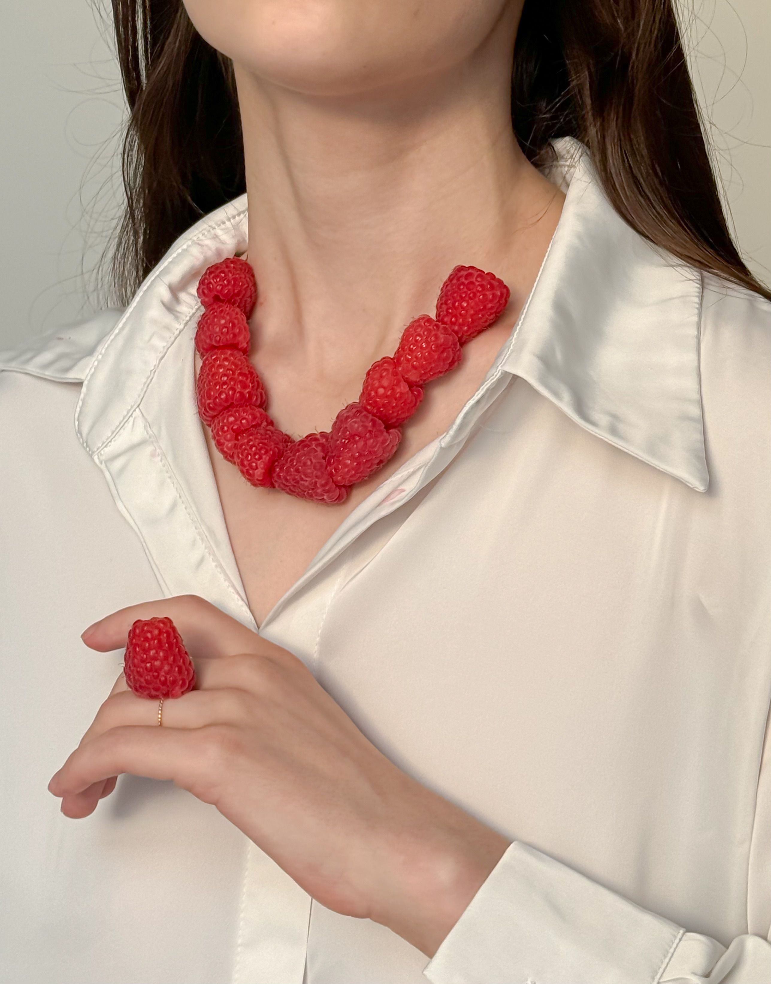 Close-up of a woman wearing a creative raspberry necklace and ring against a neutral background.