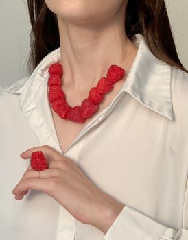 Close-up of a woman wearing a creative raspberry necklace and ring against a neutral background.