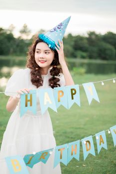 A cheerful young woman celebrates her birthday with decorations and a party hat in a sunny outdoor setting.