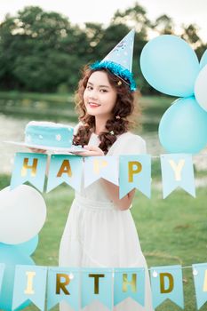 Young woman celebrating birthday outdoors with cake and balloons beside a lake.