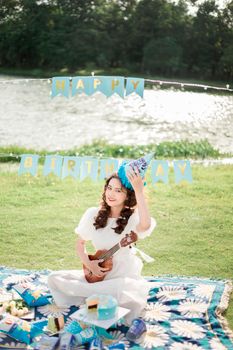 Young woman enjoys a sunny birthday outdoors, playing ukulele by the river.