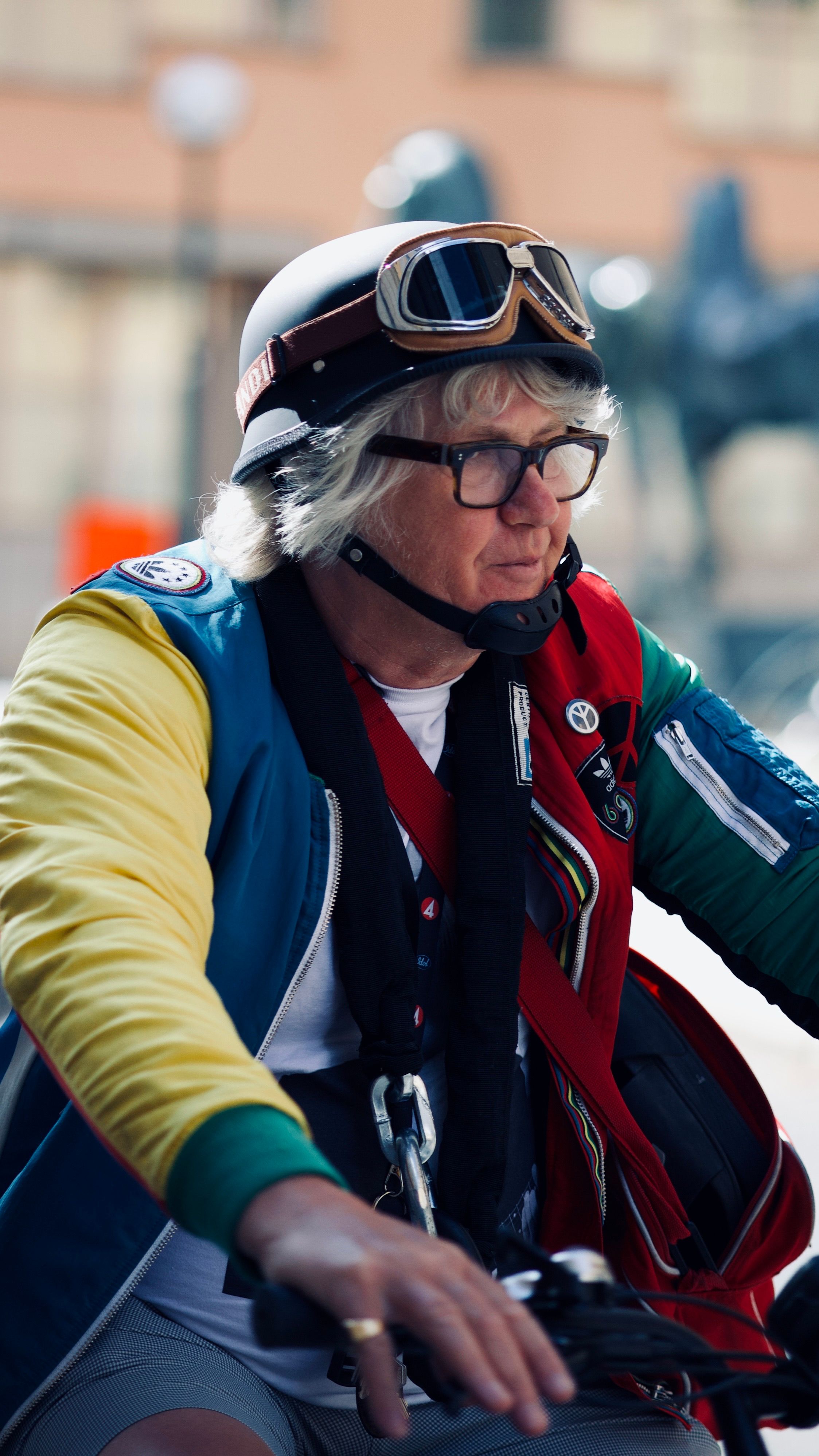 Elderly woman in vibrant jacket riding a motorcycle, wearing racing goggles.