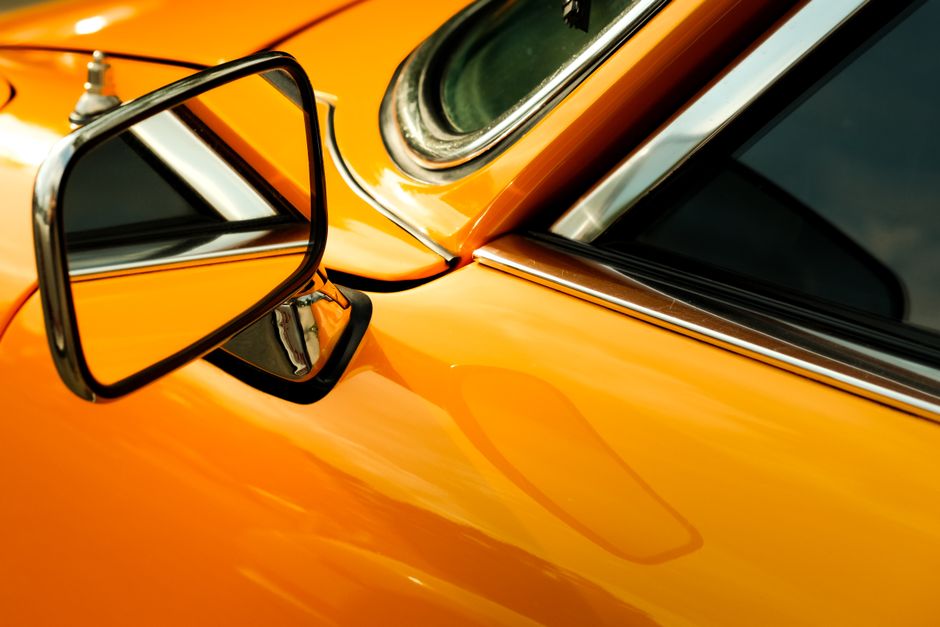 A close-up view of a vintage car's vibrant orange exterior with a chrome side mirror.