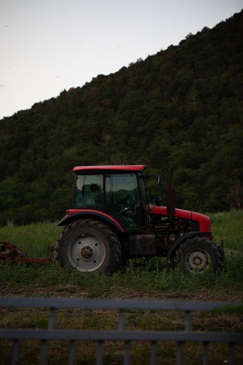Free A red tractor stands on a green field with mountains in the background, showcasing rural agriculture. Stock Photo
