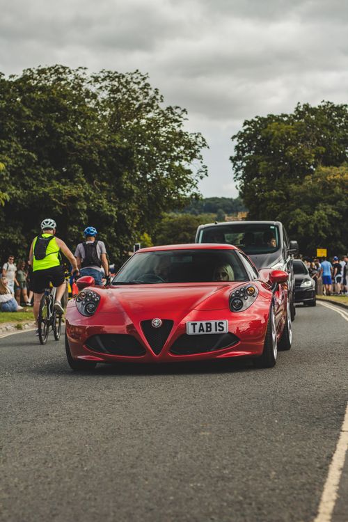 Free Red sports car on a busy road with cyclists, surrounded by greenery on a cloudy day. Stock Photo