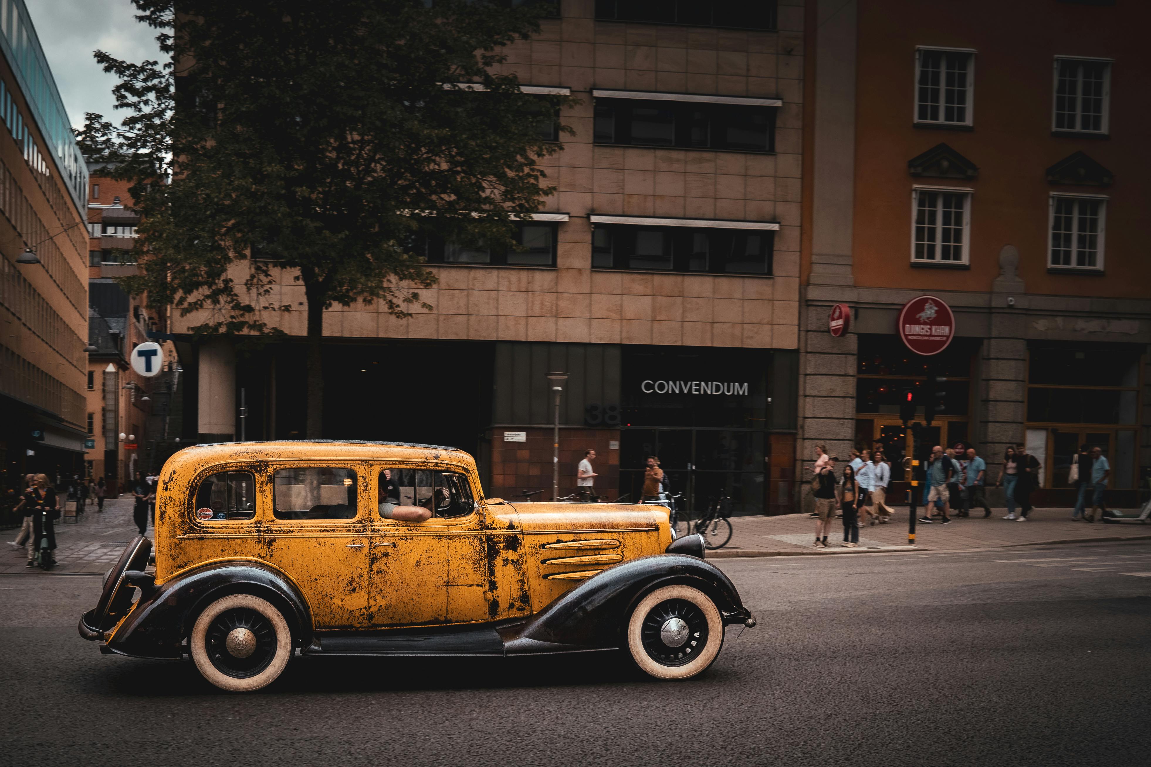 Classic yellow vintage car driving on an urban street with city architecture in the background.
