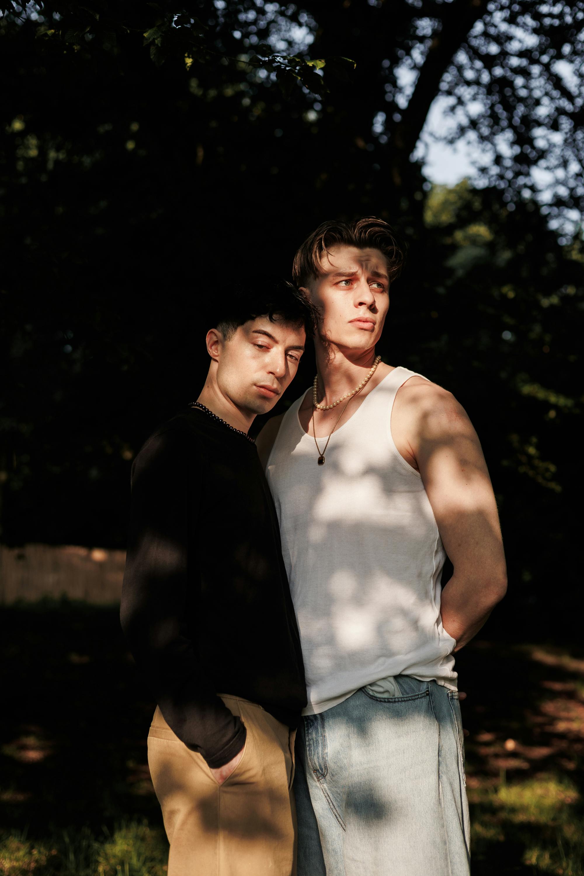 A striking portrait of two men standing in a sunlit forest, showcasing fashion and friendship.