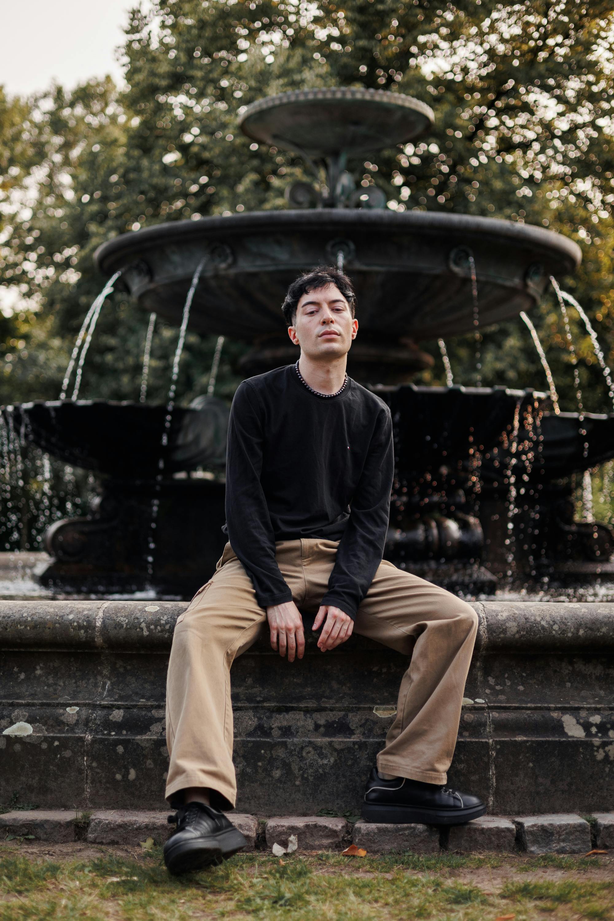 A young man sits pensively on a fountain edge in a serene park setting.