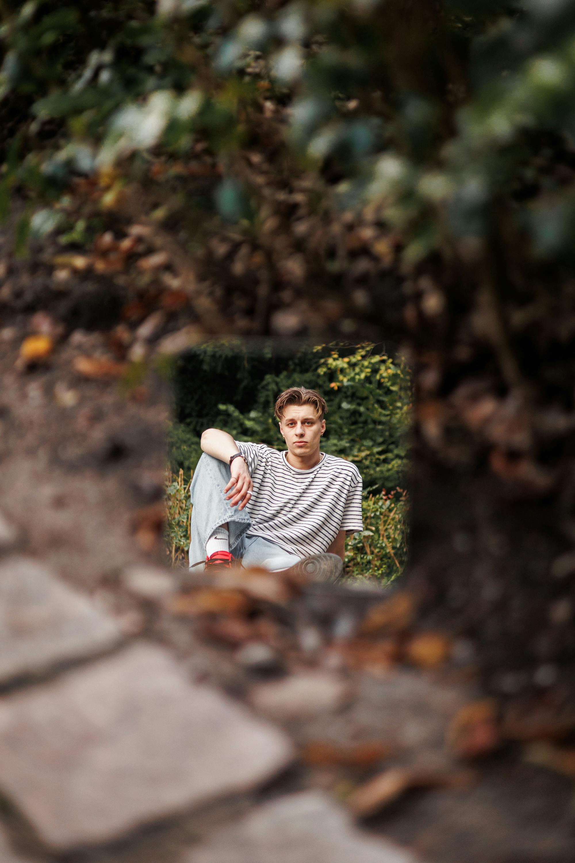 A young man sits thoughtfully in an outdoor park during autumn, surrounded by natural foliage.