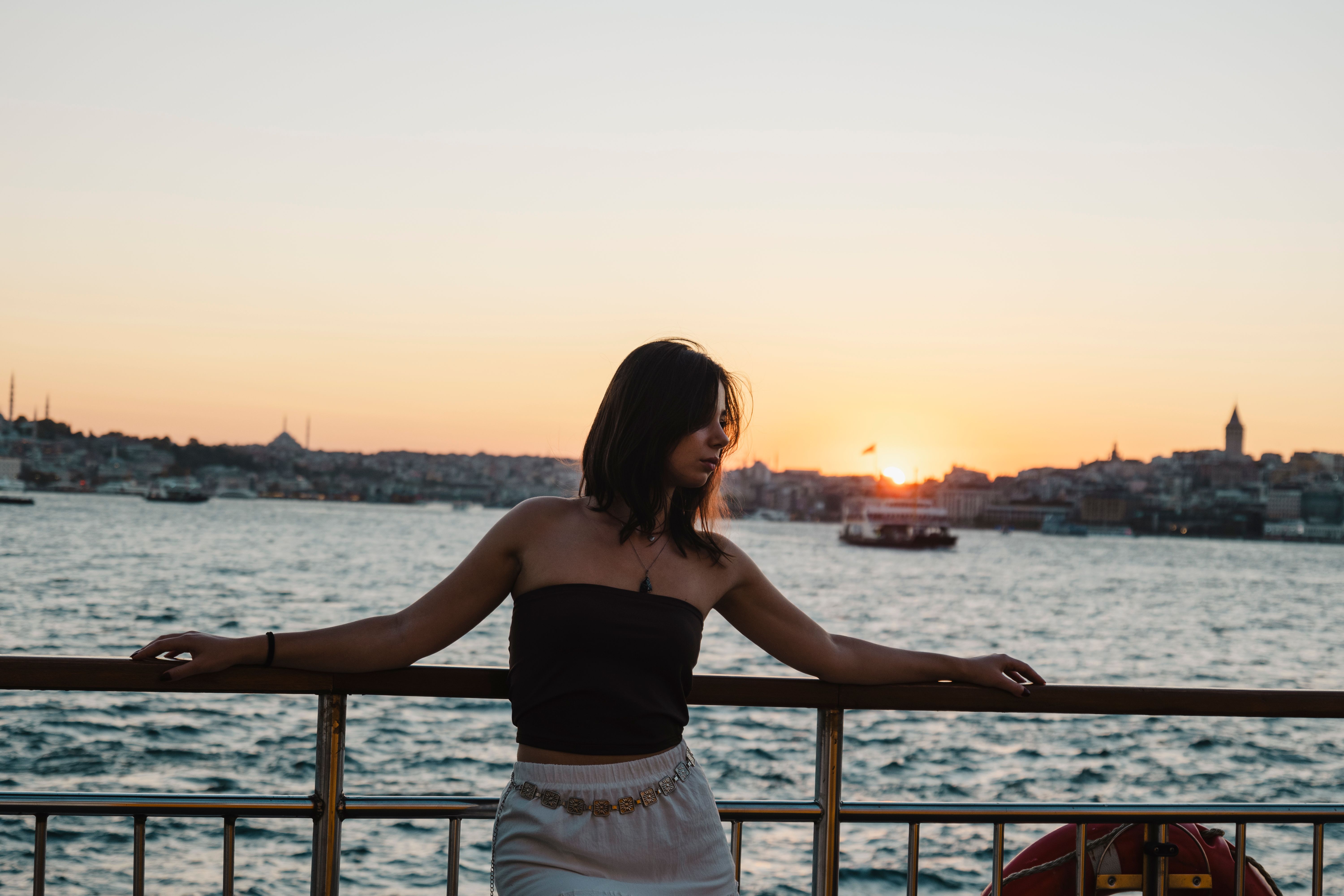 A solo female traveller stands by the Bosphorus in Istanbul, with a stunning sunset backdrop over the city skyline.