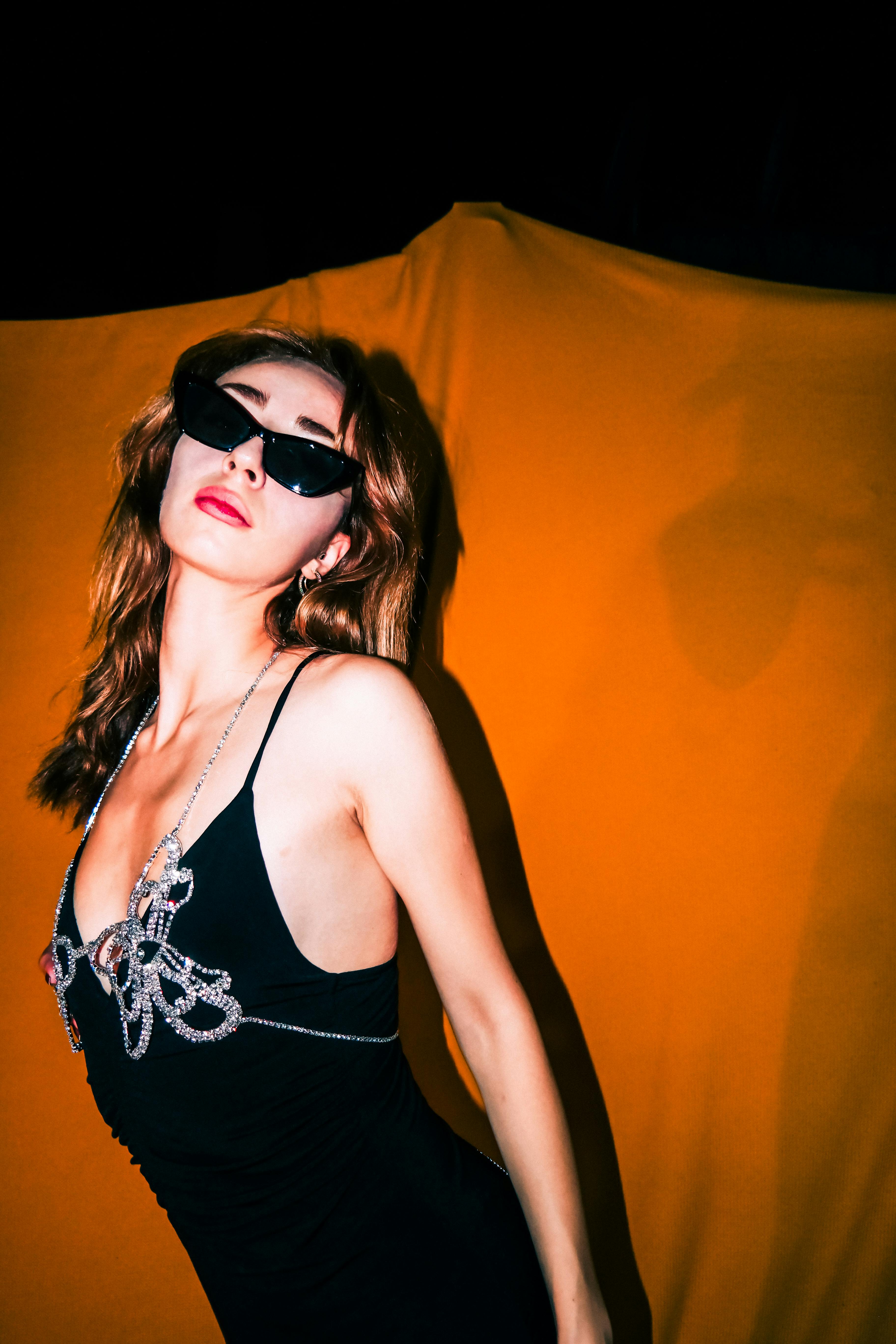 Free Fashionable woman posing in black dress with sunglasses against vibrant backdrop. Stock Photo