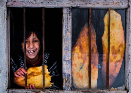 Free A joyful child holding traditional bread behind window bars, expressing warmth and culture. Stock Photo