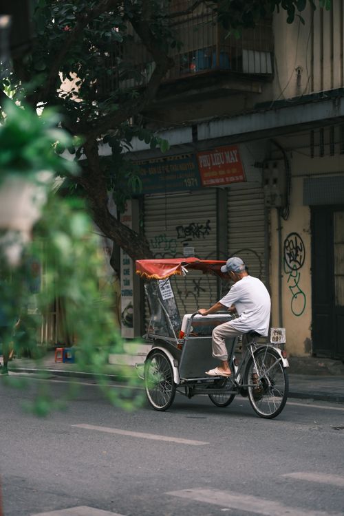 Free An elderly man riding a traditional rickshaw along a city street with urban scenery. Stock Photo