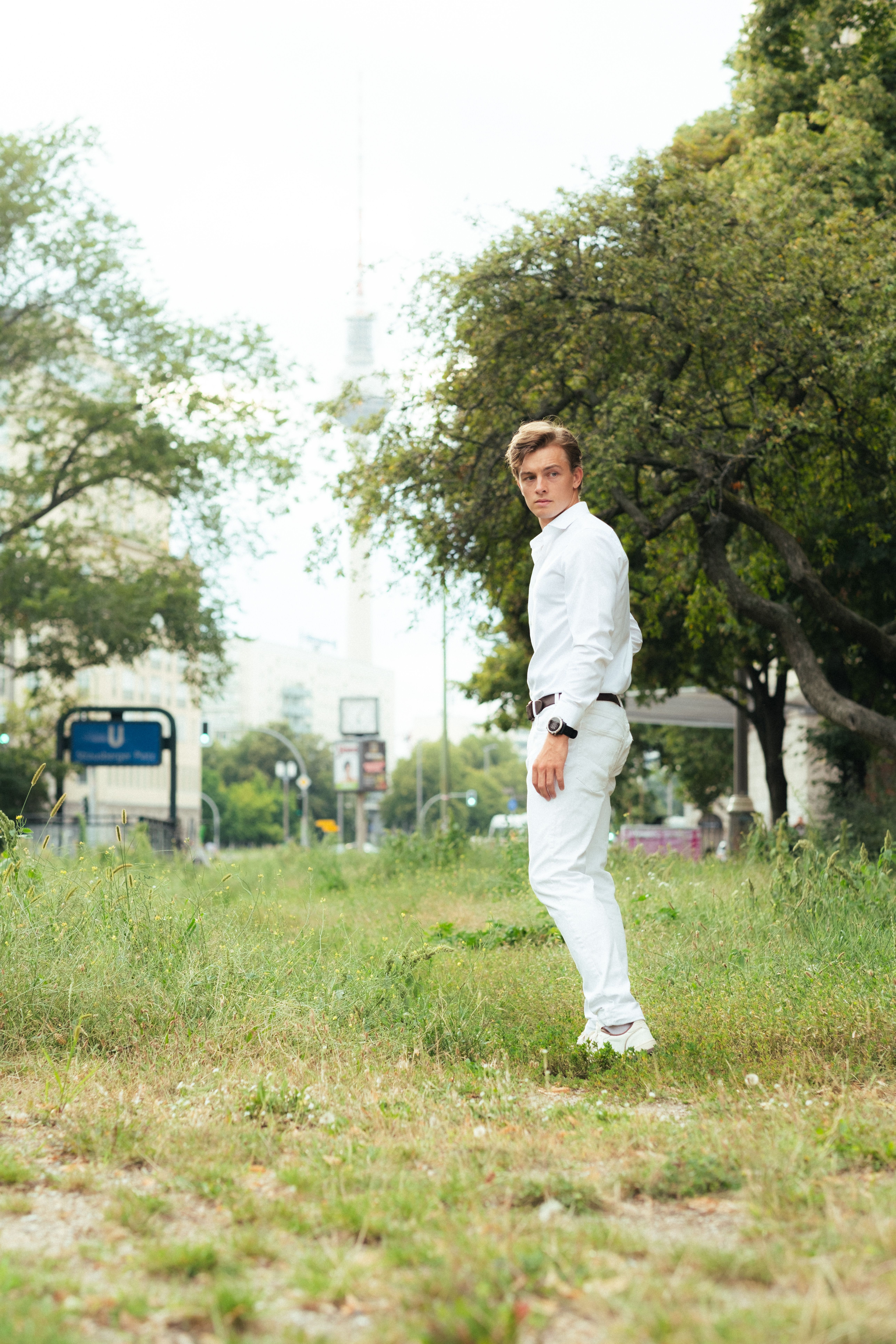 A young man in a casual white outfit standing in a Berlin park with trees and cityscape in the background.