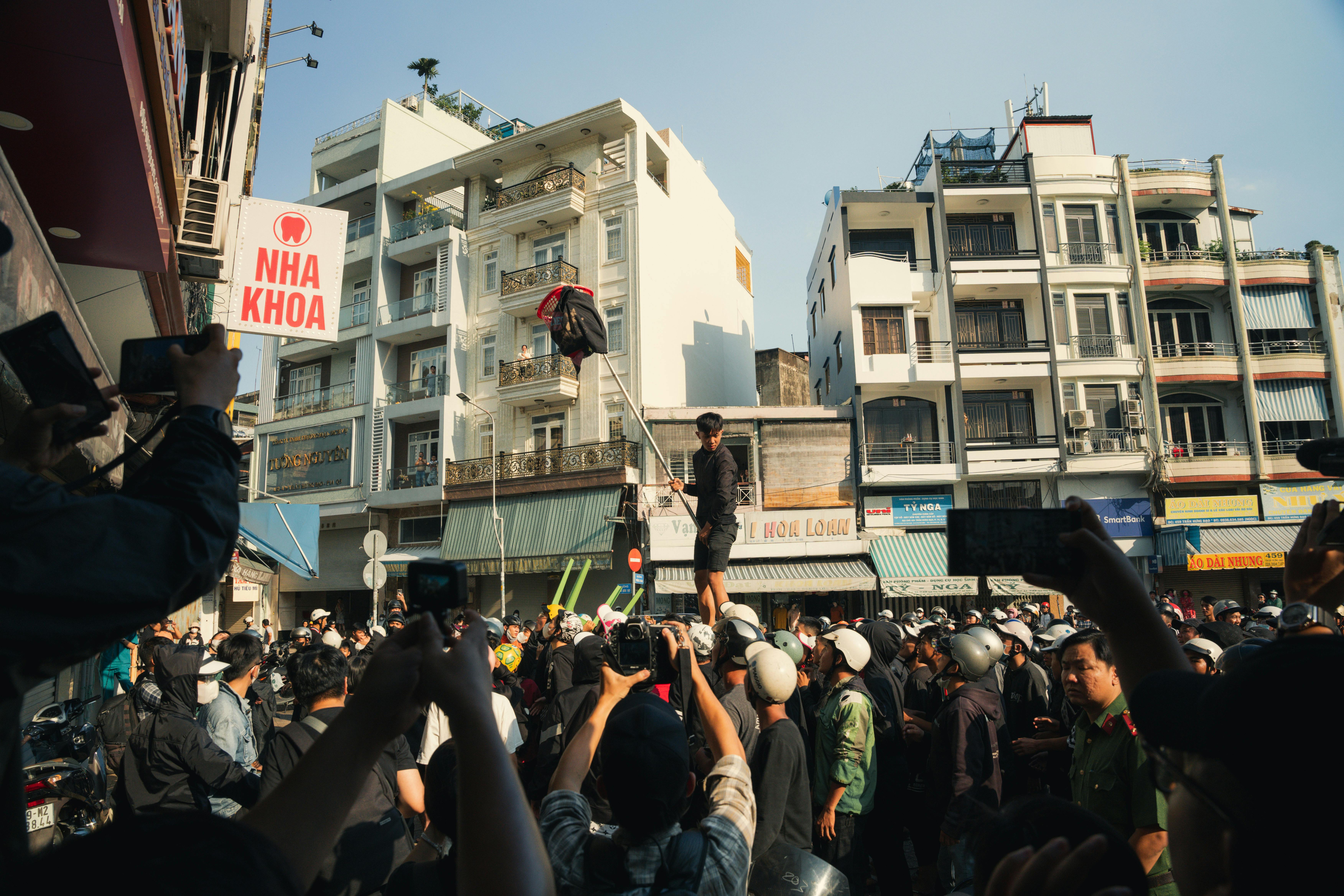 A bustling crowd gathers on a street during a lively daytime event, with buildings in the background.