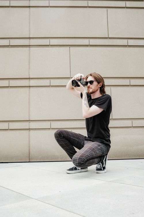 Free Young man crouching outdoors with camera by a wall, capturing urban moments. Stock Photo