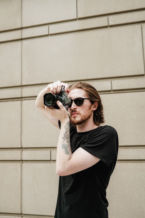 Free Casual young man wearing sunglasses, capturing urban views with a camera. Stock Photo