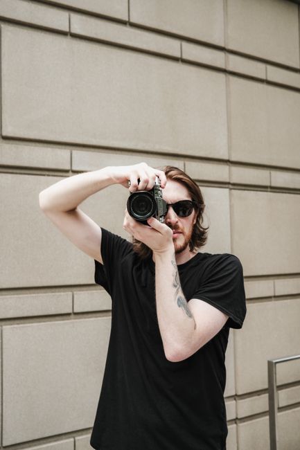 Young male photographer in urban setting using a camera, wearing sunglasses.