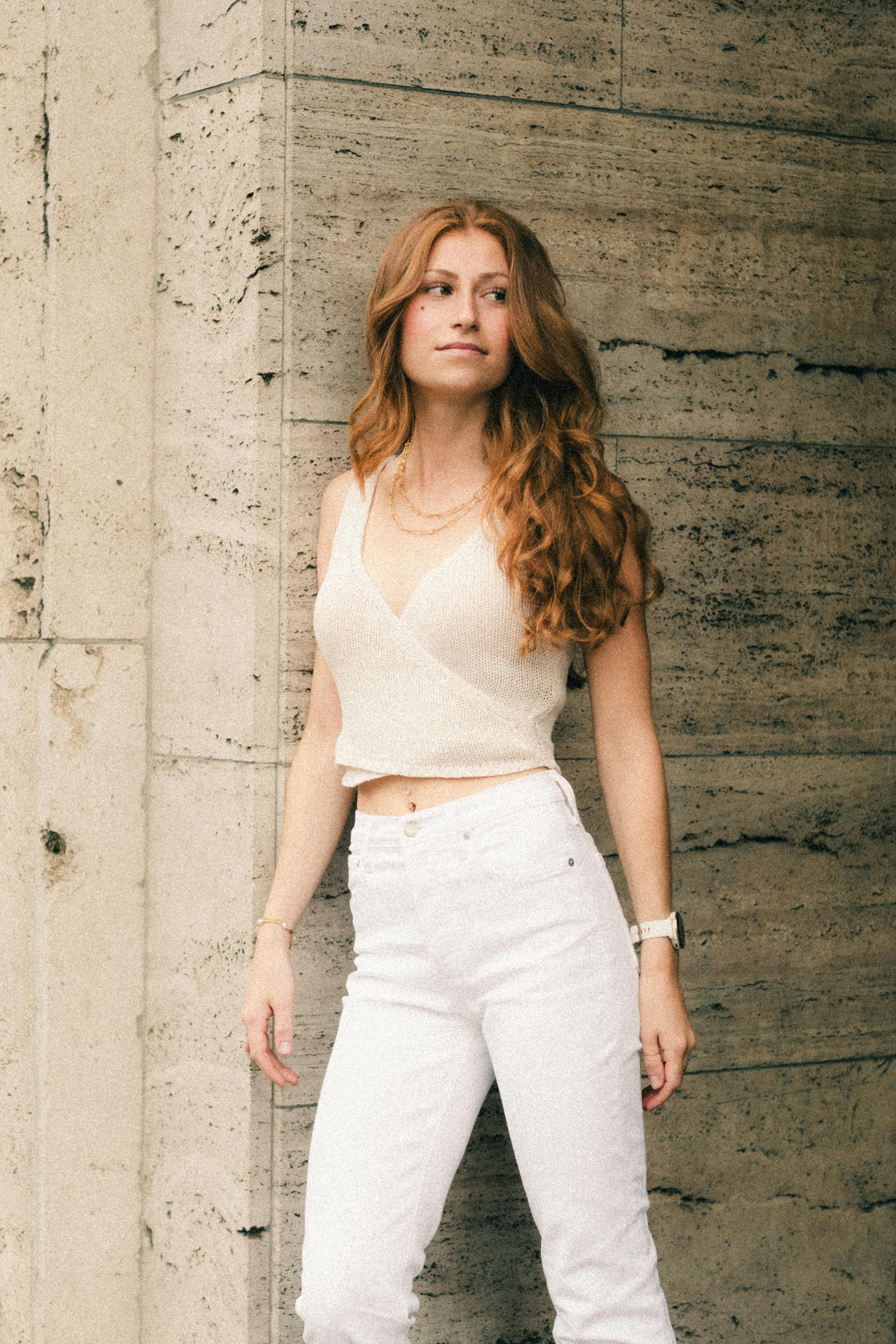Elegant young woman poses against a stone wall in Berlin wearing trendy casual attire, exuding confidence and style.
