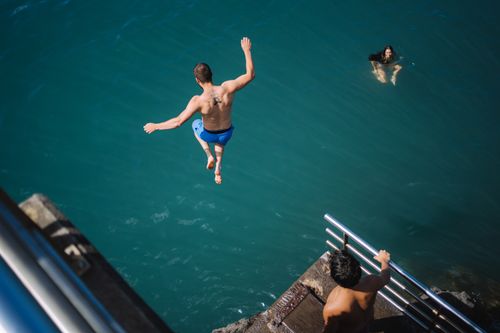 Free Man diving into the ocean from a ledge with others swimming, showcasing thrilling summer fun. Stock Photo