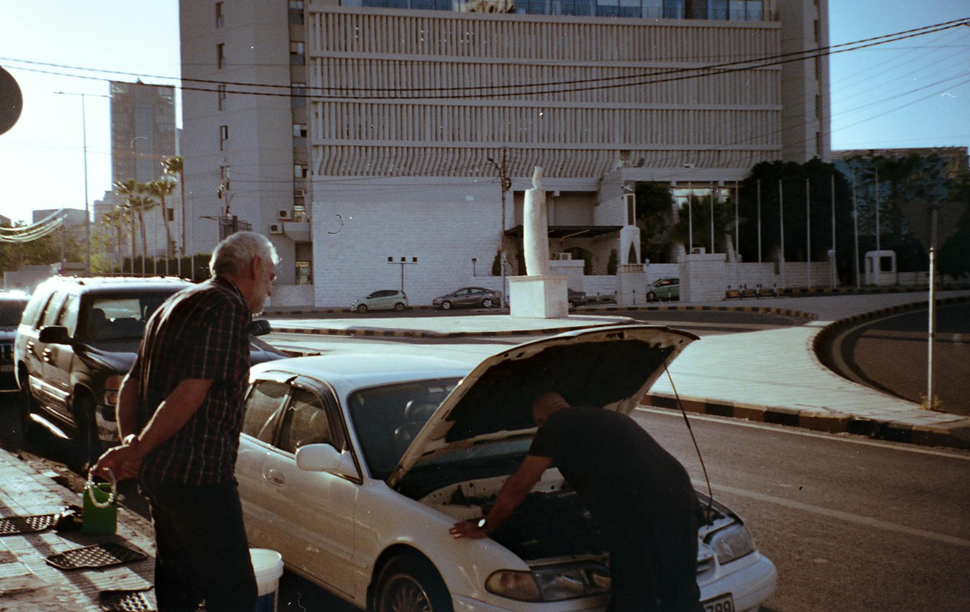 Two men repairing a car by the roadside in Amman, captured in bright daylight.