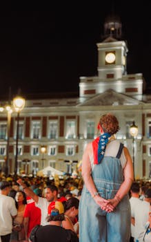 A nighttime crowd gathers under city lights near a historic building in Madrid, Spain.