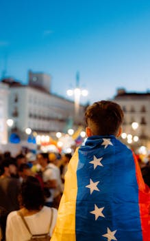 A vibrant city scene with a young man draped in the Venezuelan flag at a nighttime gathering.