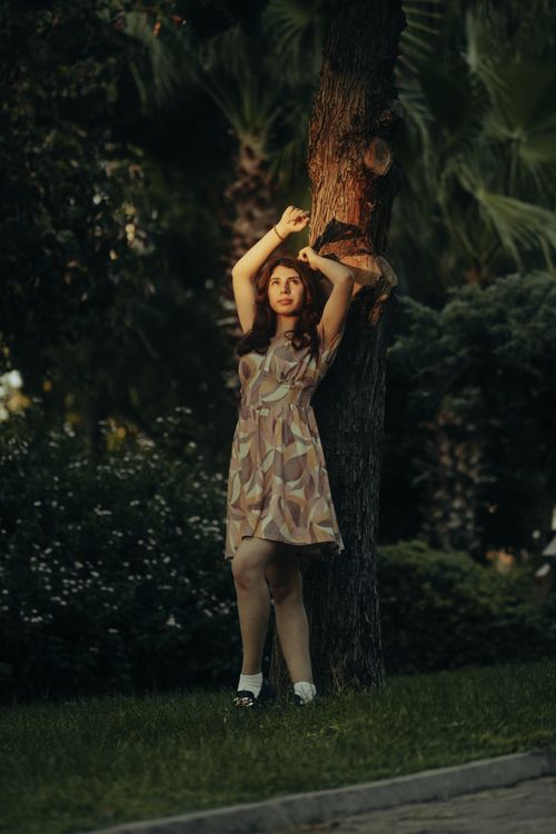 Free A woman in a patterned summer dress poses against a tree in a park setting. Stock Photo