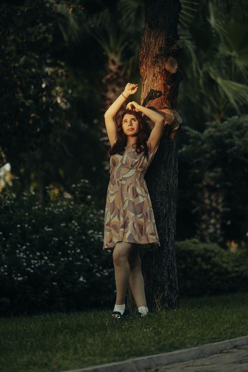 Free Glamorous woman poses in a summer dress against a tree in a park setting. Stock Photo