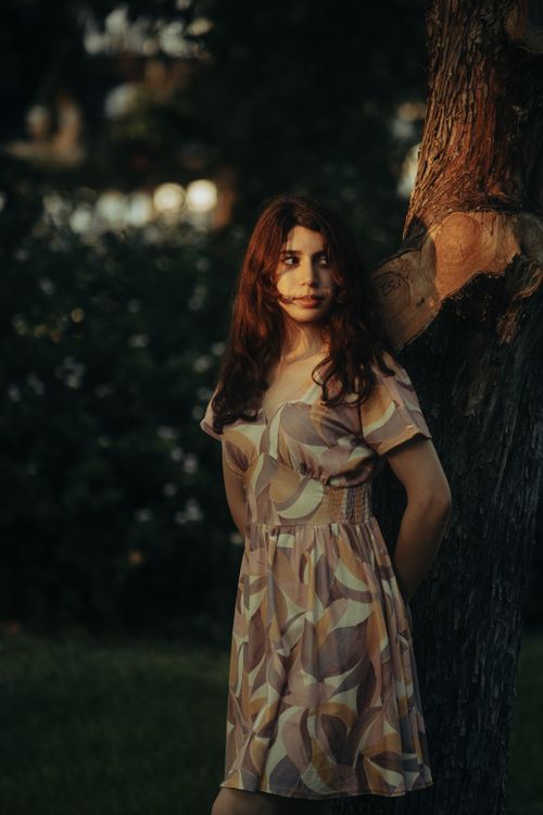 Free A young woman in a patterned dress leaning against a tree in a romantic outdoor setting. Stock Photo