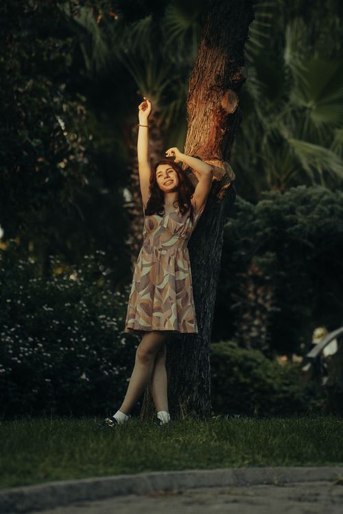 Free A woman in a patterned dress leans against a tree in a park during a warm summer evening. Stock Photo