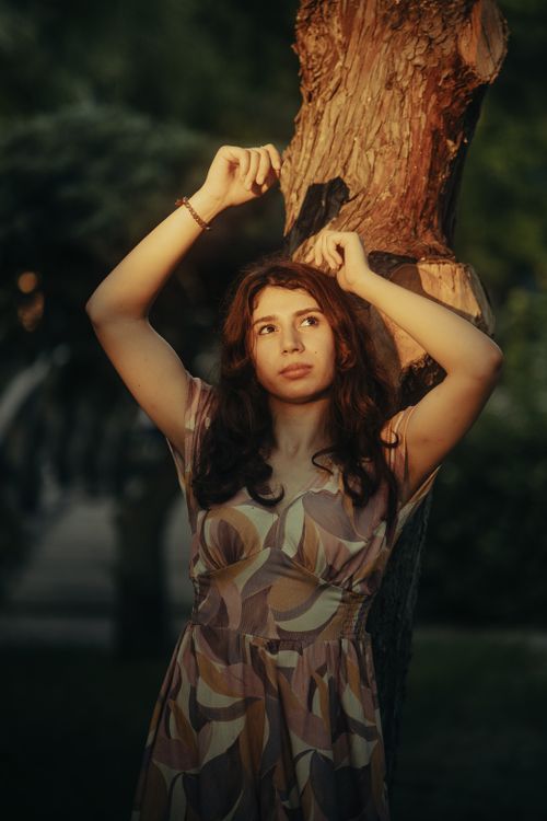 Free A beautiful woman in a patterned dress poses gracefully by a tree at sunset. Stock Photo